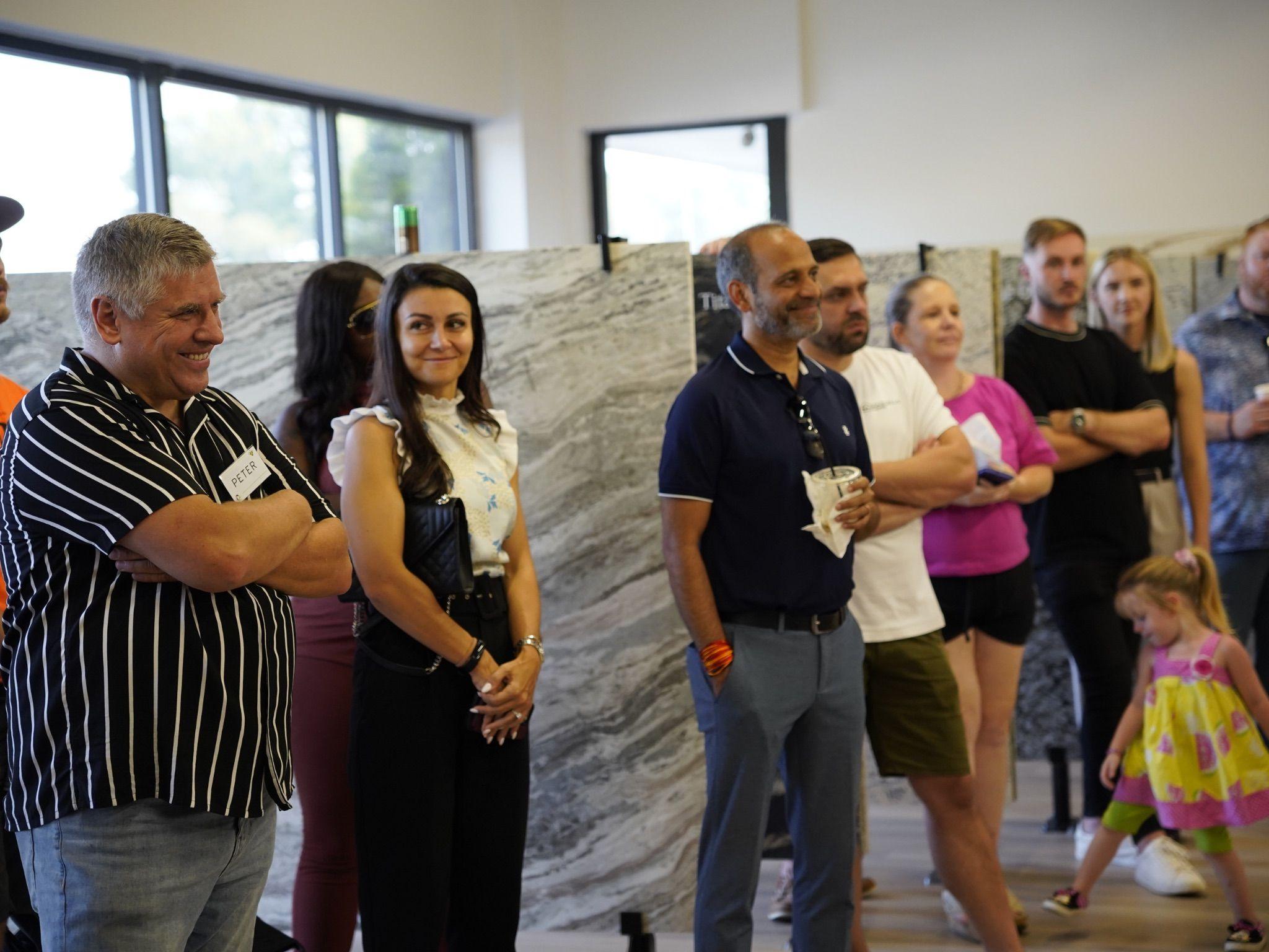 A group of attendees listens attentively during the Grand Opening celebration at the showroom, with large granite slabs displayed in the background.