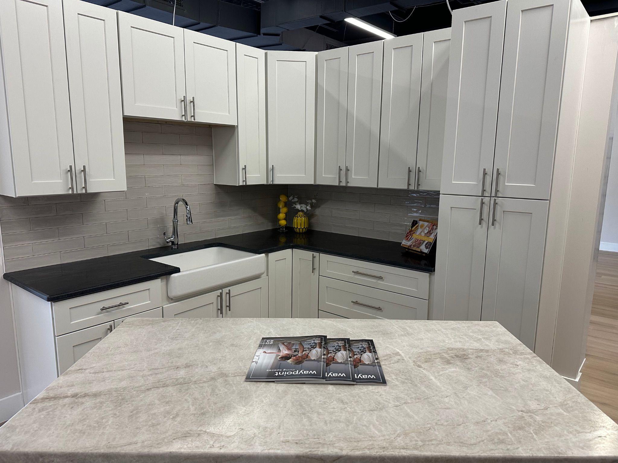 A sleek kitchen display with white cabinets, dark countertops, and a farmhouse sink, featuring a light neutral tile backsplash. A light-colored island countertop is displayed in the foreground with brochures on top.