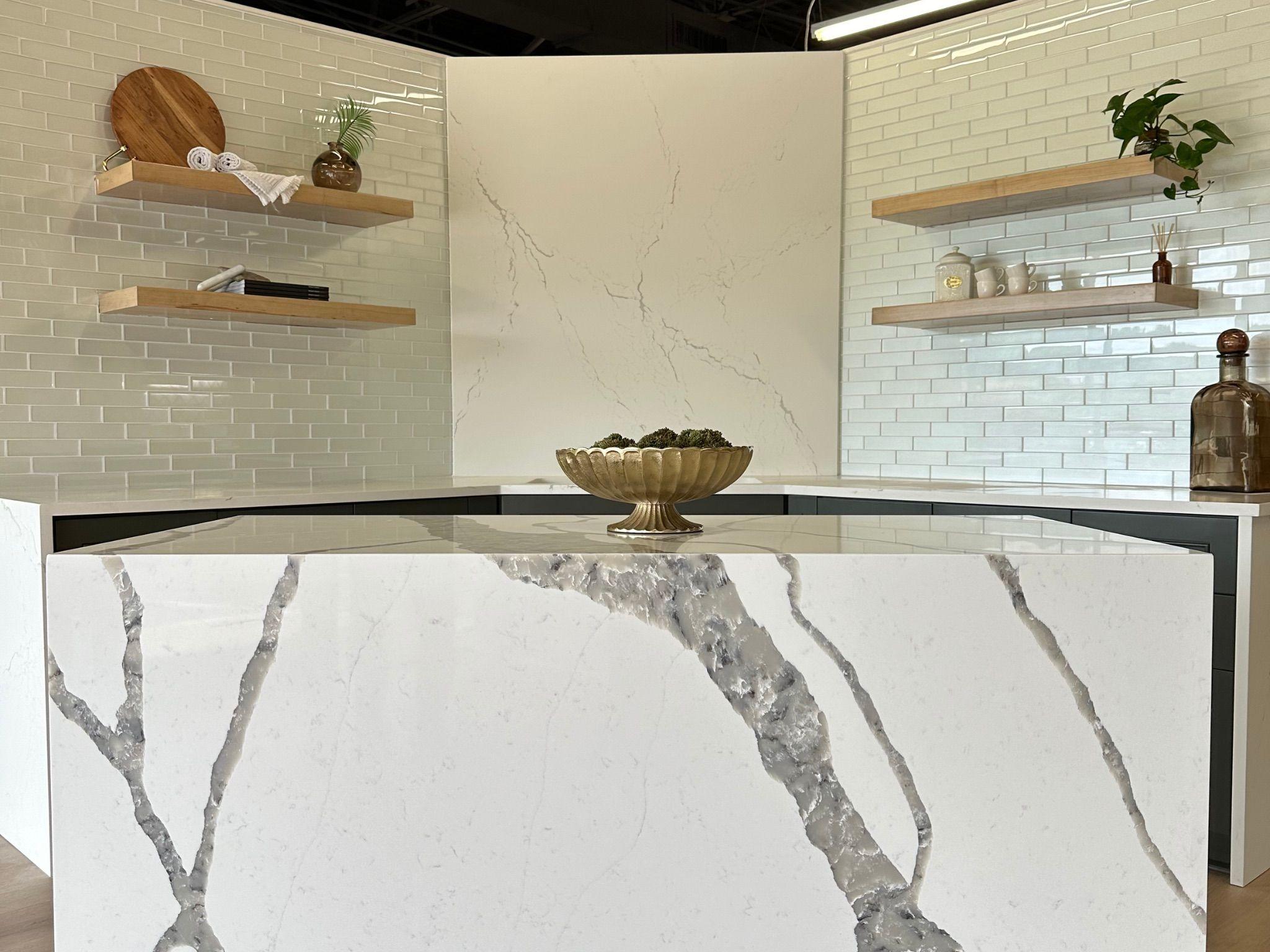 A sophisticated kitchen island with a white marble waterfall countertop featuring gray veining, set against a white tile backsplash and light wood floating shelves decorated with minimalist kitchen accents.