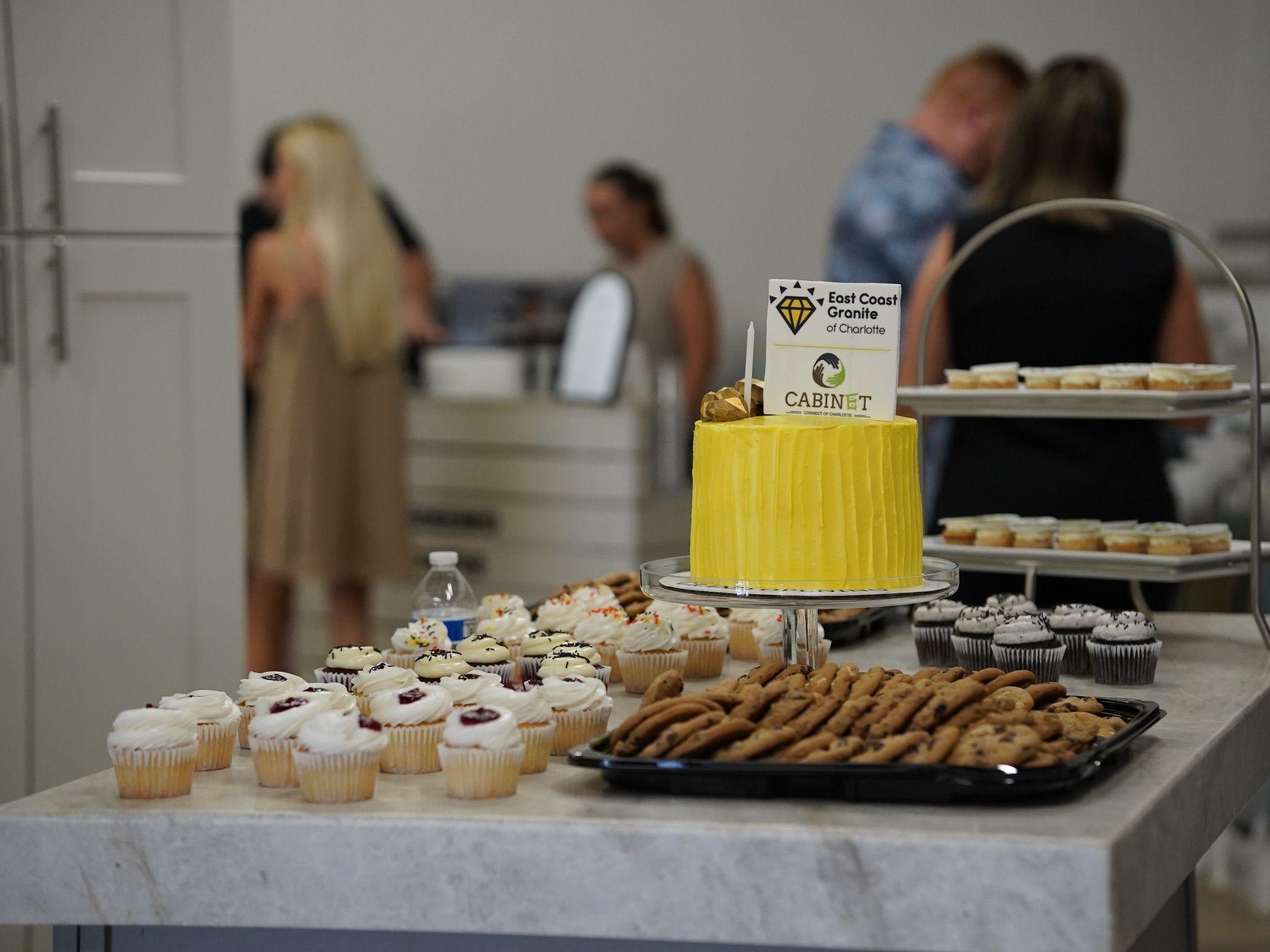 Dessert table featuring cupcakes, cookies, and a yellow cake with the East Coast Granite & Tile logo, ready to celebrate the Grand Opening event. Guests mingle in the background.