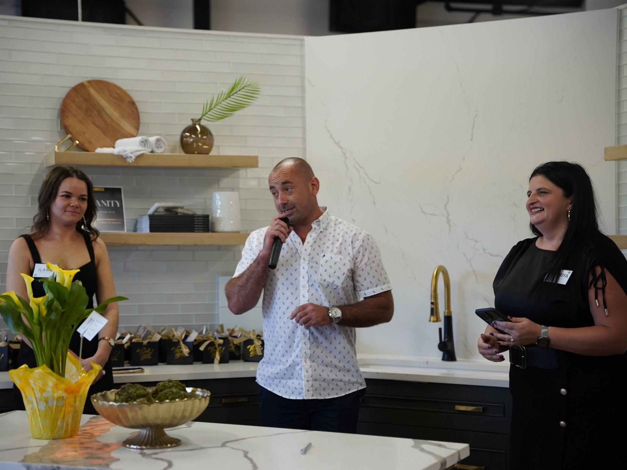 Our founder and leadership team speaking at the Grand Opening event of the East Coast Granite & Tile showroom, standing in front of a beautifully designed kitchen with a marble backsplash and gold accents.