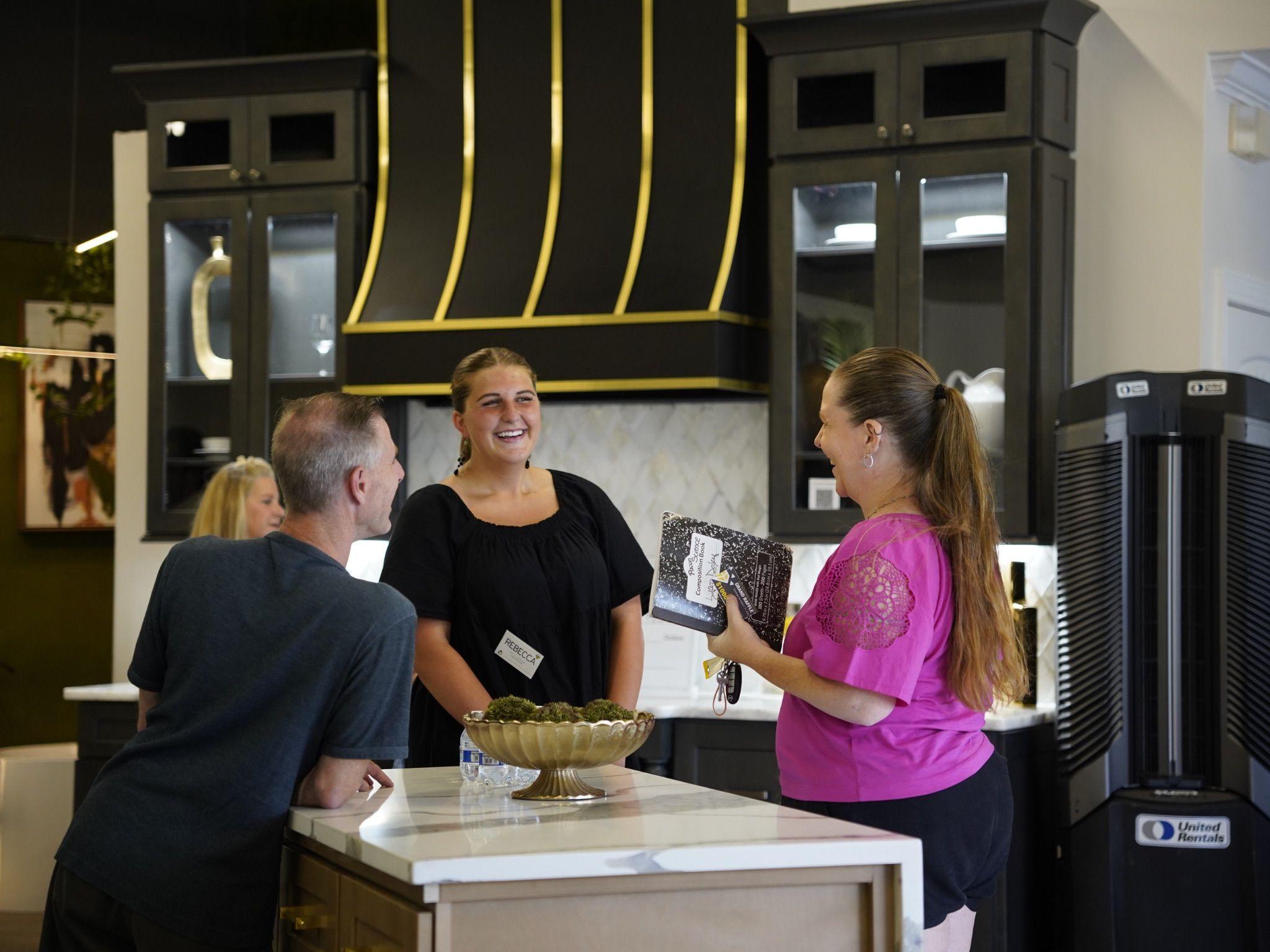 A group of guests socializing and exchanging smiles at the Grand Opening event. The modern kitchen showroom serves as the backdrop, featuring dark cabinets and a stylish vent hood with gold trim.